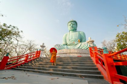 Left side view of young Asian monk hold red umbrella and walk down on stair in front of green buddha