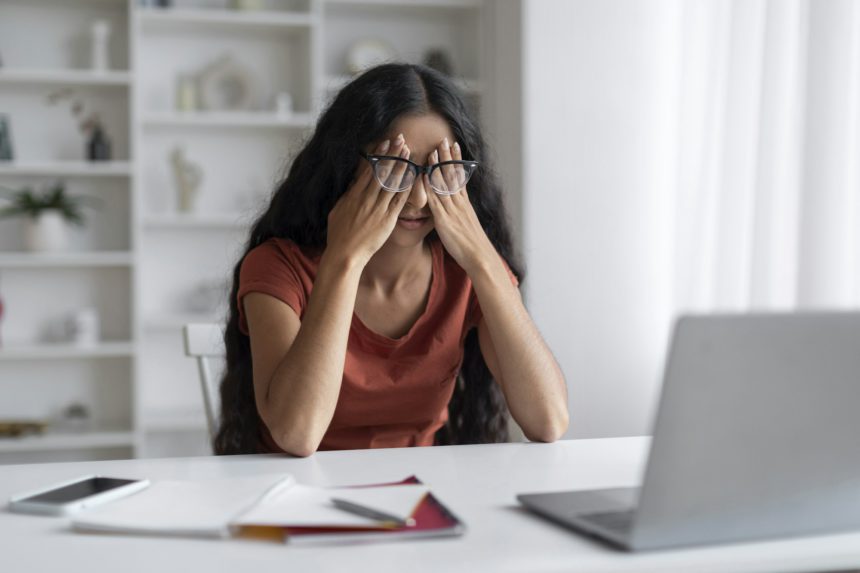 Stressed young eastern businesswoman sitting at workplace, rubbing eyes